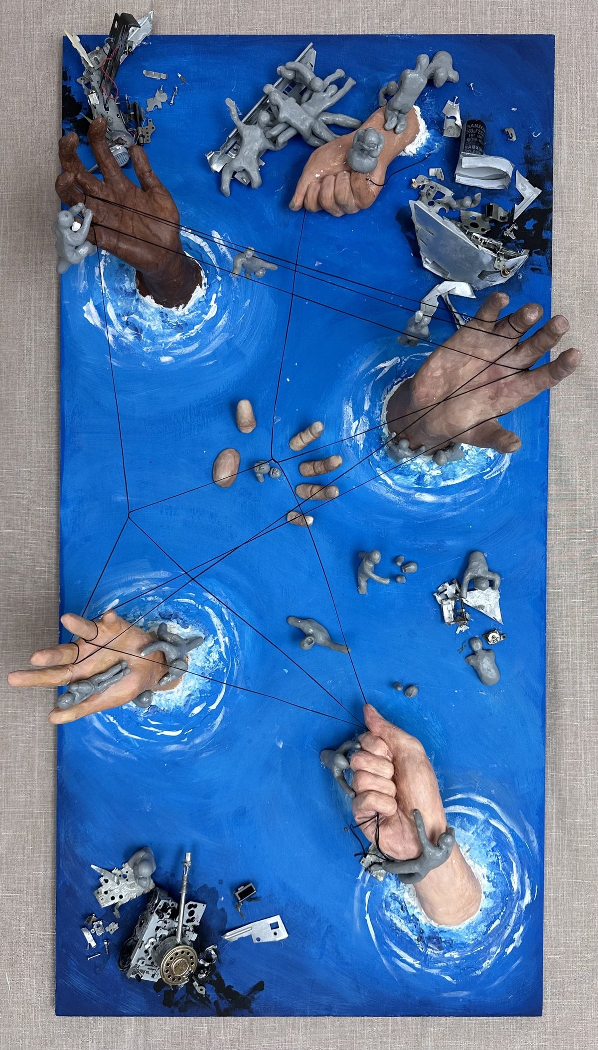 Mixed-media piece of sculpted hands reaching out of whirlpools on a blue panel, tied together with string among scrap metal